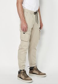 Beige Cargo-Hosen mit elastischen Bündchen, Kordelzug-Taille, mehreren Seitentaschen und einer glatten Textur. Getragen mit erdigen High-Top-Sneakern.