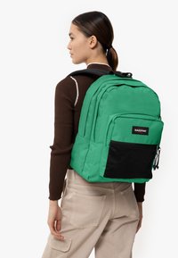 Mochila de tela verde con múltiples compartimentos, bolsillo de malla negra, correas acolchadas y un parche con logotipo. Textura suave y diseño estructurado.