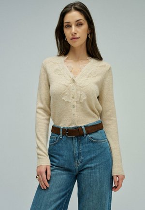 Mujer con cárdigan beige abotonado con detalles de encaje floral, jeans azules de cintura alta y cinturón marrón, de pie contra un fondo liso.