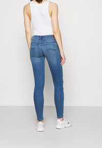 Jeans ajustados de mezclilla azul con una cintura de altura media, cinco bolsillos y un patrón de olas sutil en los bolsillos traseros. Combinados con una camiseta sin mangas blanca y zapatillas.