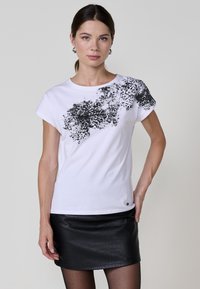 Camiseta blanca de algodón con un estampado abstracto negro en el hombro izquierdo y mangas cortas. Combinada con una falda ajustada de cuero sintético negra.