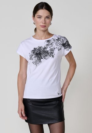 Camiseta blanca de algodón con un estampado abstracto negro en el hombro izquierdo y mangas cortas. Combinada con una falda ajustada de cuero sintético negra.