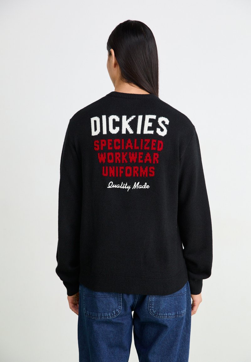 Svart stickad tröja med röd och vit text på ryggen: "DICKIES SPECIALIZED WORKWEAR UNIFORMS Kvalitetstillverkad". Ribbad ärm- och nederkant.