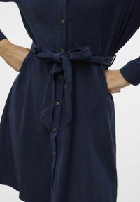 Abito in velluto a coste blu navy con bottoni frontali, maniche lunghe e cintura legata in vita. Presenta una texture liscia e un sottile motivo a coste.