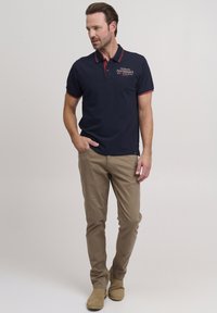 Mornarska polo majica s crvenim akcentima, ovratnikom i vezom. Uparena s tan slim-fit hlačama i bež gležnjačama. Glatka pamučna tekstura.
