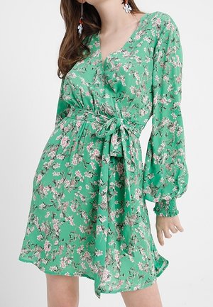 Grøn blomsterprintet slå-om-kjole med lange puffede ærmer og bindebånd i taljen, båret af en kvinde med langt hår og blomsterformede øreringe.