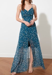 Femme portant une robe maxi à fleurs bleues avec des bretelles fines, des côtés découpés, une fente sur le devant et des sandales à talons hauts avec bride transparente, debout sur un sol en bois.