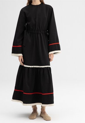 Black, long-sleeve dress with a gathered waist, featuring red zigzag accents and white fringed hem. Worn with beige woven shoes.