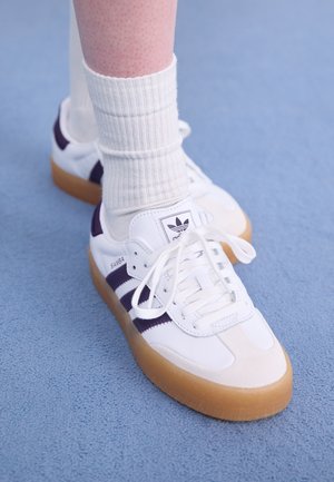 White Adidas Samba sneakers with purple stripes, featuring a rubber gum sole and laces. Worn with ribbed white socks.