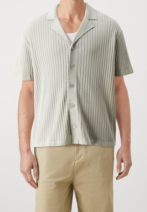 Mężczyzna w jasnoszarej, krótko rękawowej, prążkowanej koszuli na guziki, na białym podkoszulku, w beżowych spodniach, na jednolitym tle.