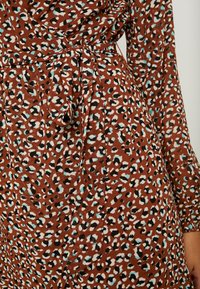 Robe portefeuille marron à imprimé léopard en noir, blanc et turquoise, avec ceinture en tissu et manches longues. Matière texturée.