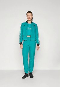 Fato de treino atlético azul-petróleo com detalhes em preto. O casaco tem riscas brancas e um logótipo; as calças têm riscas laterais e bolsos. Tênis pretos completam o conjunto.