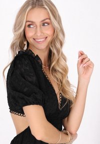 Bluză neagră scurtă cu mâneci bufante, material texturat, decolteu adânc în formă de V, ornamentată cu o bordură circulară decorativă, stilizată cu brățări suprapuse.