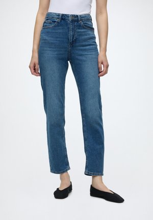 Femme portant un jean bleu taille haute et des ballerines noires, debout devant un fond clair uni.