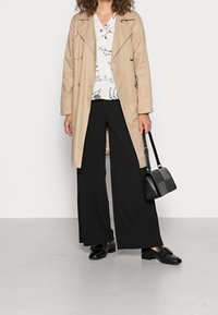 Beige trenchcoat over een witte bloemenblouse, gecombineerd met zwarte wijde broek en zwarte loafers. Een kleine zwarte handtas wordt in de hand gehouden.