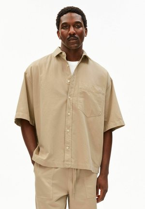 Mann trägt beige kurzärmliges Hemd mit Brusttasche und passende beige Hose, steht mit den Händen in den Taschen.