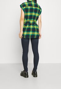 Camisa a cuadros verde y azul marino con mangas enrolladas, cintura ceñida y dobladillo curvado. Combinada con jeans oscuros y zapatos de plataforma negros.