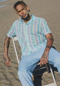 Chemise à manches courtes avec un motif bleu et blanc, associée à un pantalon bleu clair. Chaise en bois, arrière-plan de plage de sable.