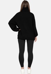 Kobieta stoi tyłem, ubrana w luźny czarny sweter z golfem, czarne legginsy i białe trampki na jednolitym tle.