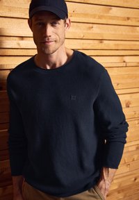 Pull en maille bleu marine foncé avec un motif texturé, col rond et détail de logo. Porté avec un pantalon beige devant un fond en bois.