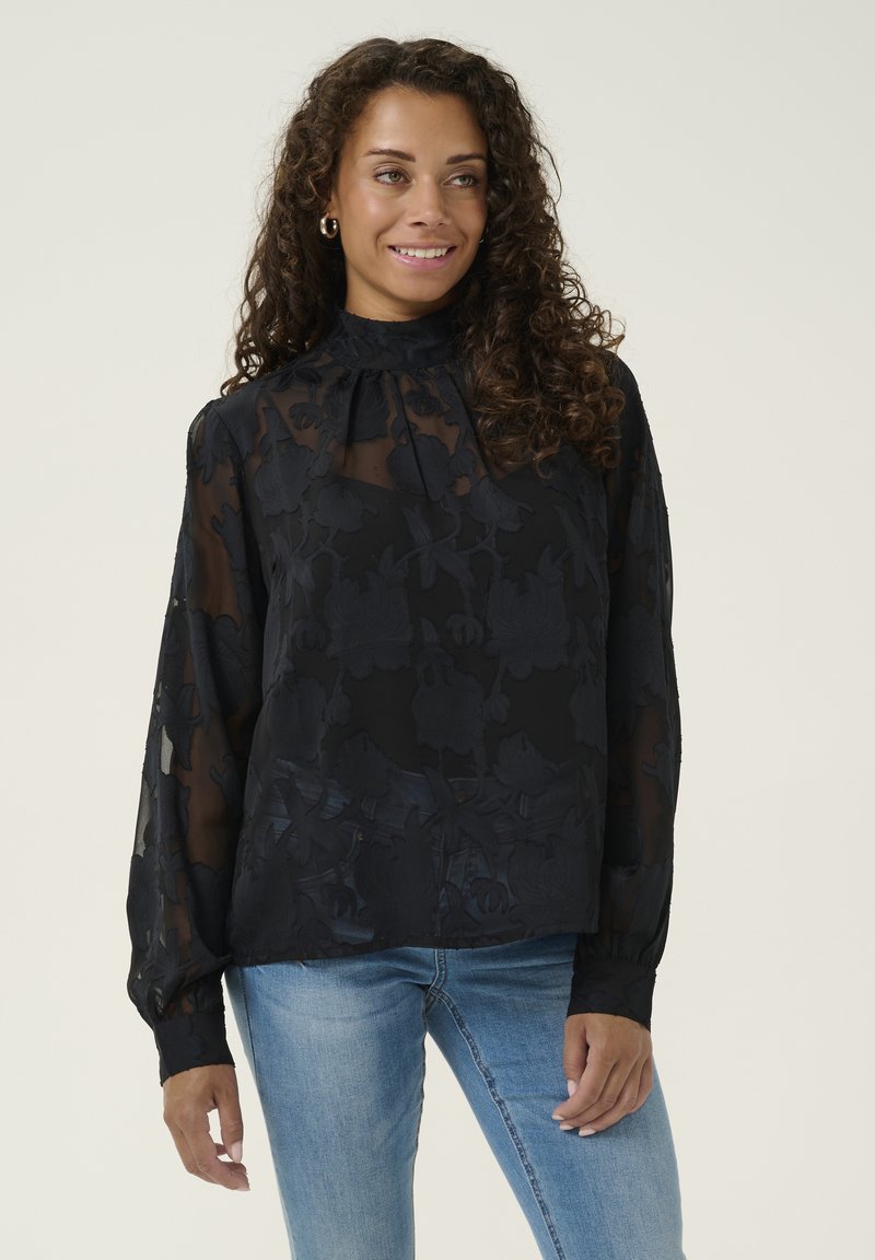 Blusa nera trasparente con motivo floreale, collo alto e dettagli arricciati. Maniche lunghe con polsini. Abbinata a jeans azzurri chiari.