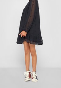 Elegant modrá šaty s dlouhými průhlednými rukávy, volánkovým lemem a jemnou vzorovanou texturou. Kombinováno s vícebarevnými vysokými teniskami.