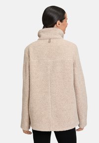 Beżowa polarowa kurtka z wysokim kołnierzem, teksturowaną powierzchnią, luźnym krojem i pętlą do wieszania. Charakteryzuje się prostym cięciem i brakiem widocznych zapięć.