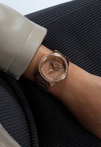 Roségoldene Uhr mit einem glitzernden, runden Zifferblatt, das ein geometrisches Design zeigt, umgeben von klaren Steinen, an einem strukturierten Metallarmband.
