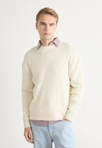 Suéter de punto acanalado color crema con un cuello redondo, llevado sobre una camisa de cuello roja y blanca a rayas. Unos jeans de mezclilla azul claro completan el look.