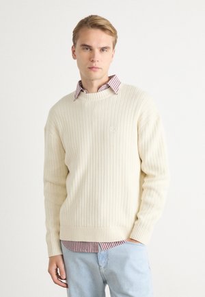 Suéter de punto acanalado color crema con un cuello redondo, llevado sobre una camisa de cuello roja y blanca a rayas. Unos jeans de mezclilla azul claro completan el look.