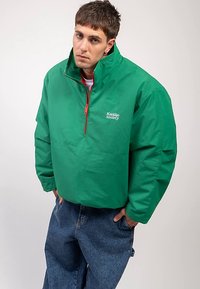 Νεαρός άνδρας που φοράει μια πράσινη μισοφ zipper ζακέτα με το κείμενο "Kaotiko Society", μπλε τζιν, στέκεται με τα χέρια στις τσέπες σε απλή φόντο.