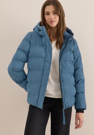 Gewatteerde blauwe jas met capuchon, voorzien van een ritssluiting en drukknopen, elastische manchetten en een gladde textuur. Draagt over een lichte top.