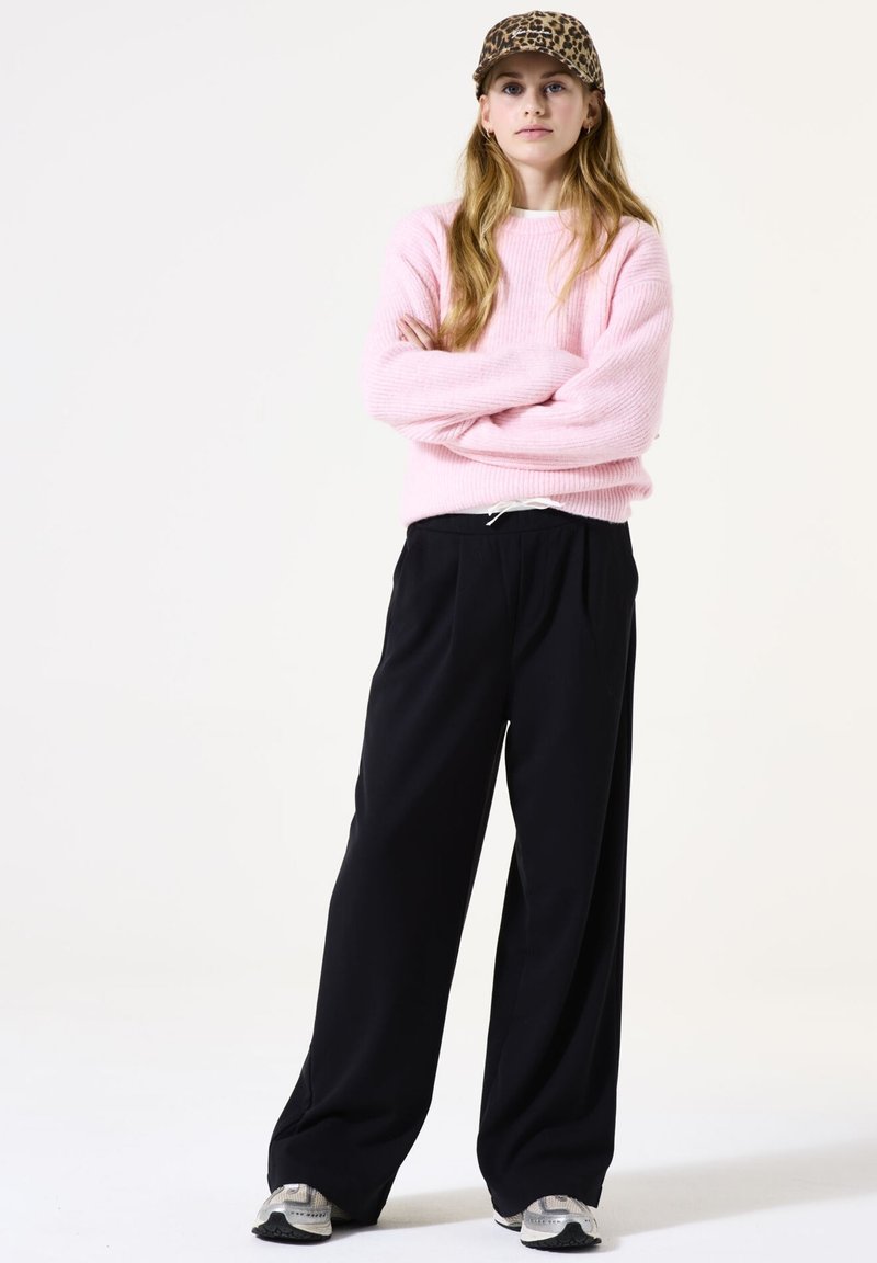 Roze geribbelde trui, zwarte wijde broek, en grijze sneakers. De hoed heeft een luipaardprint. Model staat met zijn armen over elkaar tegen een plain achtergrond.
