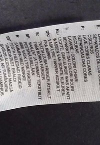 Etykieta pielęgnacyjna odzieży z instrukcjami prania ciemnych kolorów osobno, napisana w kilku językach europejskich na białym materiale.