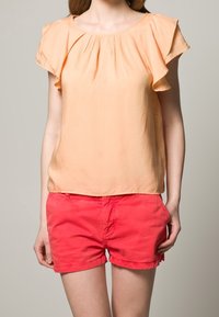 Persikofärgad blus med korta volangärmar, samlad hals och rosa shorts med fransad fåll, tillverkad av lättviktsmaterial för komfort.