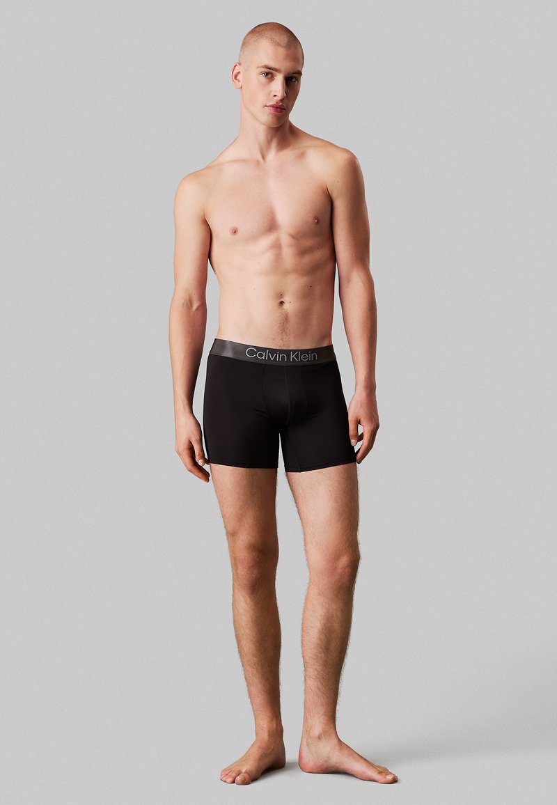 Svart boxer shorts med en tettsittende design og glatt tekstur, med en grå Calvin Klein-logo på den elastiske linningen.