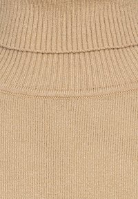 Beige ribbad stickad tröja med hög, vikbar krage. Texturen är mjuk och elastisk, med ett jämnt och konsekvent mönster genom hela plagget.