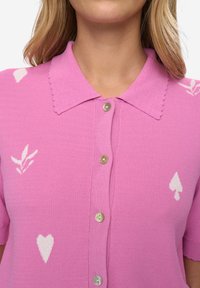 Růžová pletená polokošile s krátkými rukávy, vroubkovaným límečkem a bílými vzory srdcí a listů. Zapínání na šest knoflíků vpředu.
