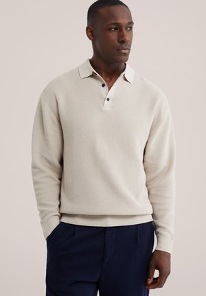 Mann trägt ein beiges, langärmliges, strukturiertes Poloshirt und dunkelblaue Hose, schaut nach rechts und hat die Hände teilweise in den Taschen.