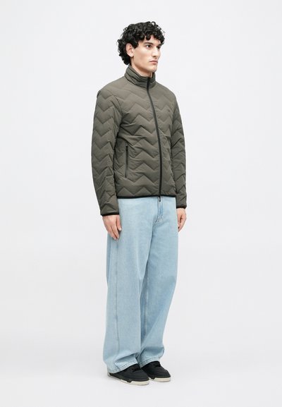 Tmavozelená olivová prešívaná bunda s vysokým golierom, vybavená predným zipsom a dvoma bočnými vreckami, v kombinácii so svetlomodrými džínsami s širokými nohavicami.