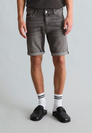 Man met opgerolde vervaagde zwarte spijkershorts, witte sokken met zwarte strepen en zwarte instapschoenen, staand tegen een effen achtergrond.