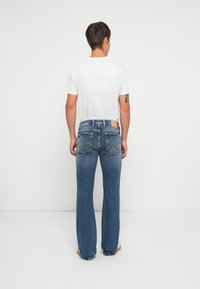 Mann stående med ryggen til, iført en hvit t-skjorte og blå rette jeans med synlige baklommer og en tatovering på venstre arm.