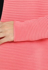 Różowy ribbowany materiał ukazuje zbliżenie długiego rękawa bluzki. Tekstura jest miękka, z poziomymi prążkami i dopasowanym miejscem na nadgarstek.