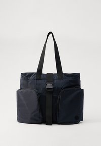 Sac fourre-tout bleu marine avec une finition en nylon texturé, deux poches latérales zippées et une bandoulière adjustable maintenue par une boucle en plastique.