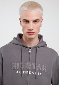 Grå zip-hoodie med "BIG STAR AUTHENTIC" trykt i hvid og grå. Har snoretræk, en frontlomme og en blød tekstur.