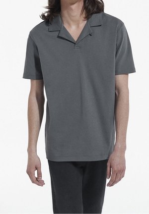 Personne portant un polo uni gris foncé à manches courtes et un pantalon noir sur fond blanc.
