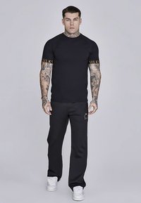 Zwarte getailleerde T-shirt met gouden "SILKS" tekst op de mouwen, gecombineerd met zwarte losse broek en witte sneakers. Gladde stoftextuur.