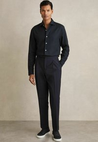 Donkerblauwe blouse met lange mouwen, kraag en witte knopen, gecombineerd met op maat gemaakte marineblauwe broek en zwarte sneakers op een neutrale achtergrond.