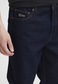 Vaqueros de mezclilla azul oscuro con costuras de cobre, que presentan una pequeña etiqueta de marca en el bolsillo delantero. Textura suave, diseño de pierna recta.