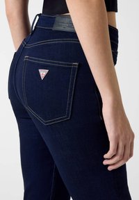 Σκούρο μπλε τζιν με απαλό υφασμάτινο φινίρισμα, πίσω τσέπη με τριγωνικό κόκκινο λογότυπο και κίτρινη ραφή κατά μήκος των ραφών.